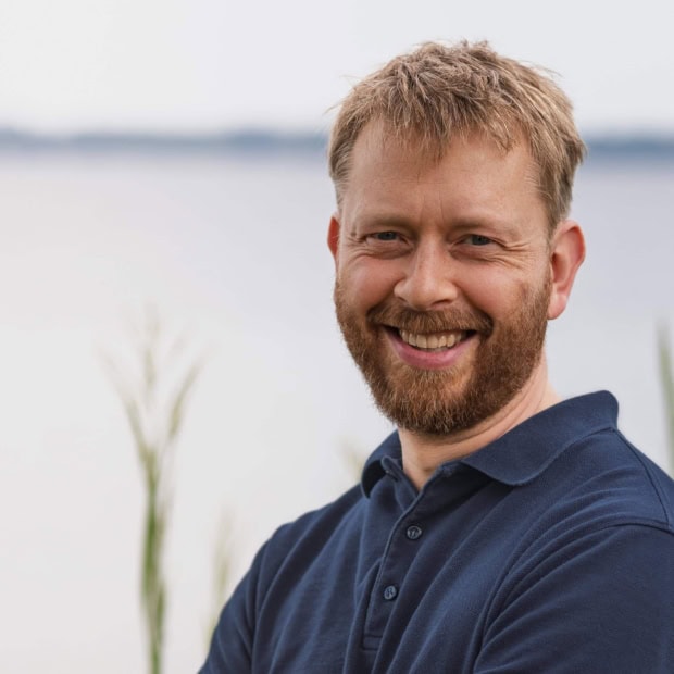 01. Alexis Philipp Lächelnder Mann vor Wasser mit Natur im Hintergrund.