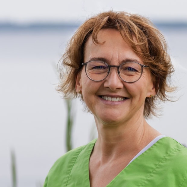 05. Helga Stricker Ärztin in grüner Praxisuniform vor Wasser mit Natur im Hintergrund.
