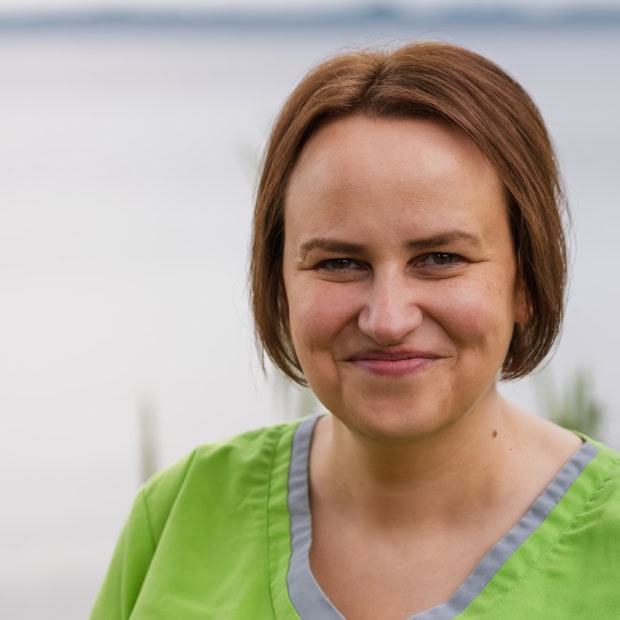 06. Jennifer Kamphaus Ärztin in grüner Klinikuniform vor Wasser mit Bäumen im Hintergrund.